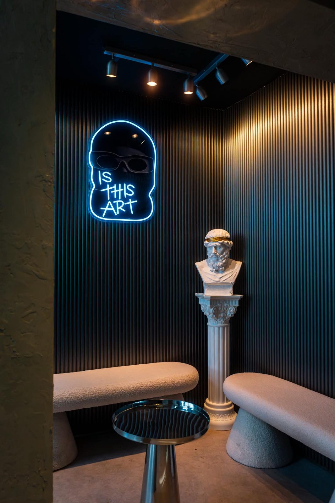 The waiting corner at Culture Box: a cyan IS THIS ART neon on black fluted panelling, a marble bust in a gold laurel on a fluted column, and two bouclé benches.